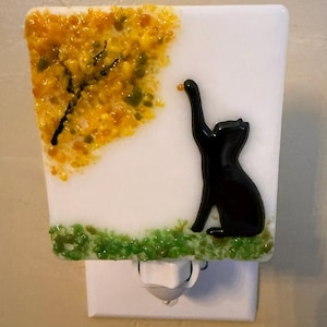 May include: A fused glass night light featuring a white rectangular base with a black cat silhouette reaching toward a textured yellow and orange tree canopy in the upper left corner and a patch of green glass frit grass along the bottom edge.
