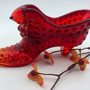 May include: A red glass shoe with a textured surface and a high heel. The shoe is a decorative item and is made in the shape of a traditional women's shoe.