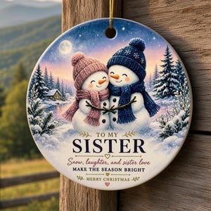 May include: A round ceramic Christmas ornament featuring two snowmen wearing pink and blue knit hats and scarves. The text reads: TO MY SISTER, Snow, laughter, and sister love, MAKE THE SEASON BRIGHT, MERRY CHRISTMAS. It hangs from a gold string.