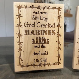 May include: A rectangular wooden plaque with engraved text and a barbed wire border. The text reads "And on the 8th Day God Created MARINES and the devil said Oh Shit!" The plaque features a light wood color.