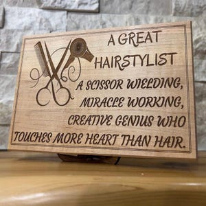 May include: A wooden plaque with engraved text and hairstyling tools. The text reads "A GREAT HAIRSTYLIST, A SCISSOR WIELDING, MIRACLE WORKING, CREATIVE GENIUS WHO TOUCHES MORE HEART THAN HAIR."