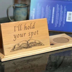 May include: Wooden book holder with the text "I'll hold your spot" and an engraved open book design. The holder has a rectangular base with a circular coaster. A blue book and a mug are in the background.