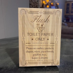 May include: A rectangular wooden sign with engraved text and decorative borders. The sign reads "Flush TOILET PAPER ONLY" and includes a list of items not to flush. The sign is made of light-colored wood.
