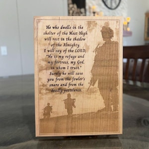 May include: A wooden plaque with a laser-etched design featuring a soldier silhouette and a religious quote. The text reads, "He who dwells in the shelter of the Most High will rest in the shadow of the Almighty."