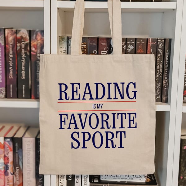 May include: A cream-colored tote bag with the text "READING IS MY FAVORITE SPORT" in navy blue and red. The bag is hanging in front of a white bookshelf filled with books of various colors and sizes.