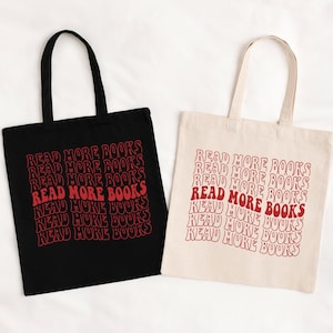 May include: Two tote bags, one black and one cream, with the text "READ MORE BOOKS" in a repeating, wavy red font. The bags have matching handles and are likely made of canvas or a similar material.