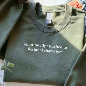 Może przedstawiać: Zielona bluza z nadrukiem "emocjonalnie przywiązany do fikcyjnych postaci" w kolorze białym na przodzie.