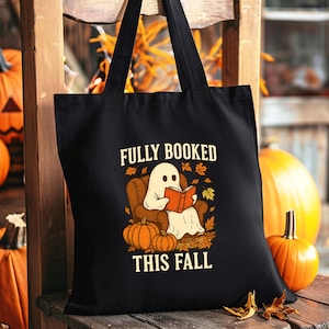 Könnte beinhalten: Schwarze Tragetasche mit dem Text "FULLY BOOKED THIS FALL" und einer Grafik eines Geistes, der ein Buch liest. Das Design umfasst Kürbisse und Herbstblätter. Die Tasche hat schwarze Griffe und wird auf einem Holzstuhl präsentiert.