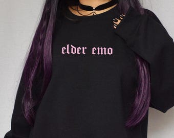 Può includere: Felpa nera con la scritta "elder emo" stampata in rosa sul davanti. La modella indossa un choker nero.