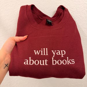May include: A maroon sweatshirt with the phrase "will yap about books" embroidered in white. The sweatshirt is folded and held by a hand with a small cross tattoo. The garment is made of a soft material.