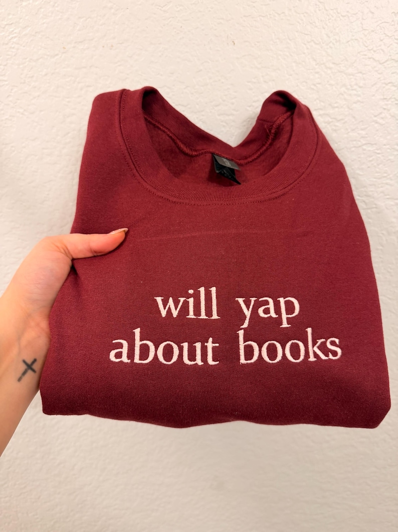May include: A maroon sweatshirt with the phrase "will yap about books" embroidered in white. The sweatshirt is folded and held by a hand with a small cross tattoo. The garment is made of a soft material.