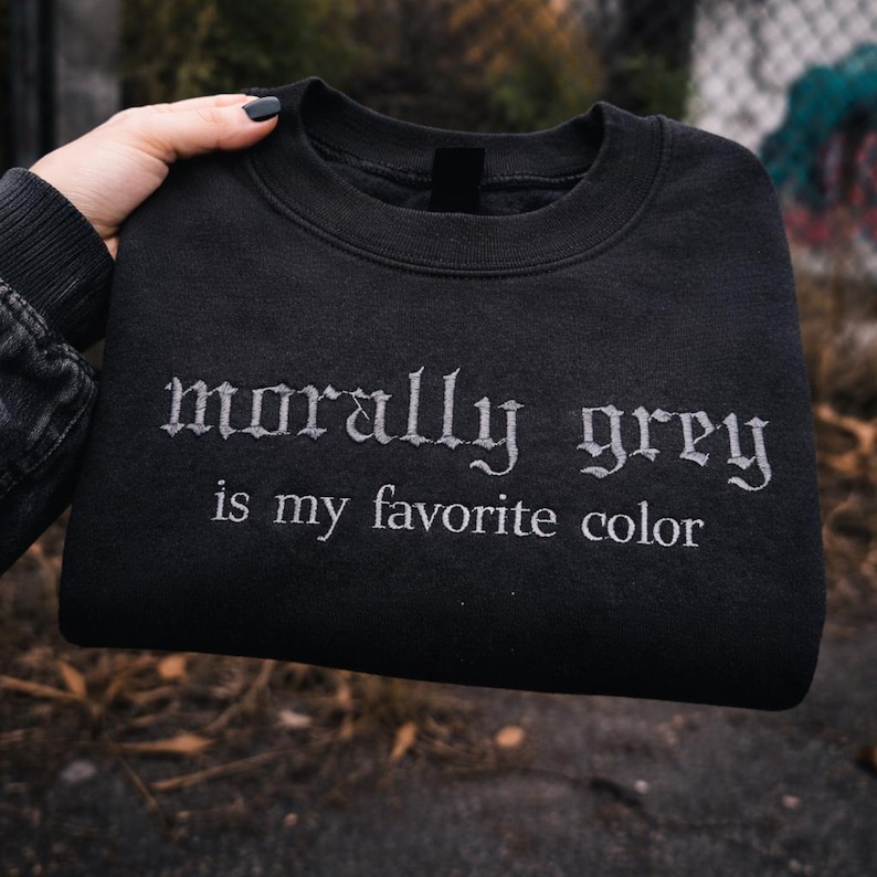 May include: A black sweatshirt with the embroidered text "morally grey is my favorite color" in a gothic font. The sweatshirt is held up by a hand, showcasing the design against a blurred outdoor background.