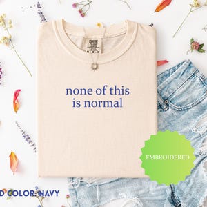 May include: A cream-coloured t-shirt with the phrase "none of this is normal" in navy blue. The shirt is embroidered and is shown with denim shorts and floral decorations. The thread colour is navy.
