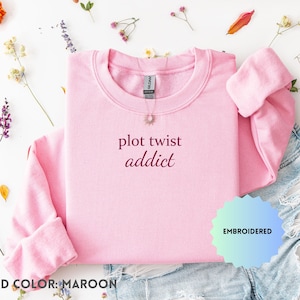 May include: A light pink sweatshirt with the words "plot twist addict" embroidered in maroon thread. The sweatshirt is displayed with denim shorts and floral accents, with the word "EMBROIDERED" in a blue and green design.