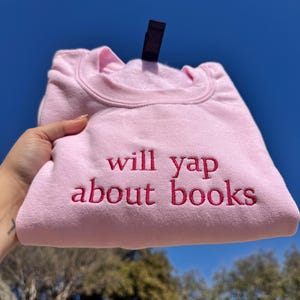 May include: A light pink sweatshirt with the embroidered text "will yap about books" in a darker pink. The sweatshirt is folded and held up against a blue sky.