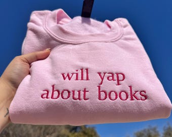 May include: A light pink sweatshirt with the embroidered text "will yap about books" in a darker pink. The sweatshirt is folded and held up against a blue sky.