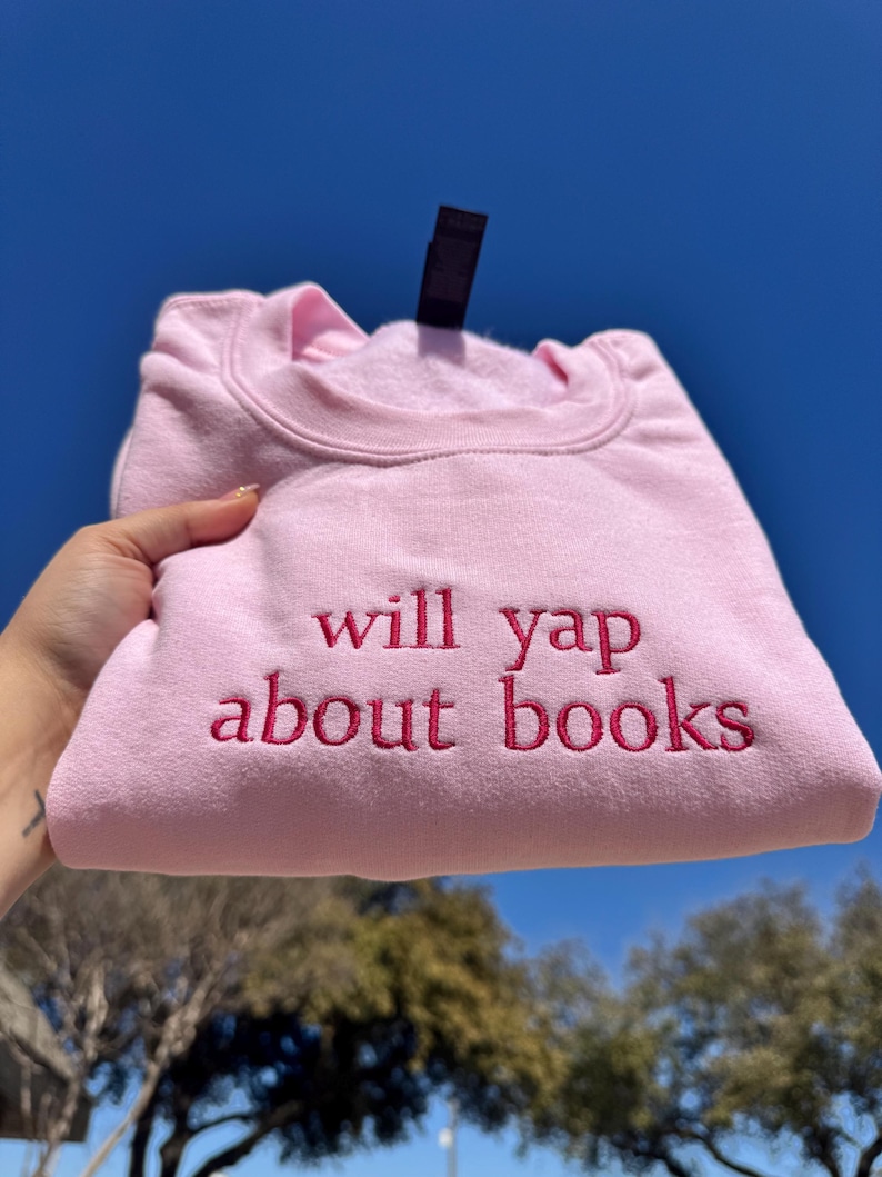 May include: A light pink sweatshirt with the embroidered text "will yap about books" in a darker pink. The sweatshirt is folded and held up against a blue sky.