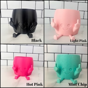 K&ouml;nnte beinhalten: Vier Keramik-Blument&ouml;pfe in verschiedenen Farben: Schwarz, Hellrosa, Pink und Mintchip. Jeder Topf ist in Form einer Cartoonfigur mit zwei Armen, die in einem Mittelfinger-Gestus erhoben sind, und zwei Beinen. Die T&ouml;pfe haben ein Smiley-Gesicht mit zwei Augen und einem Mund.