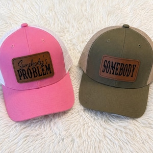 May include: Two trucker hats with mesh backs. One hat is bright pink with a white mesh back and a brown leather patch reading Somebody's PROBLEM. The other hat is olive green with a tan mesh back and a brown leather patch reading SOMEBODY.