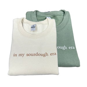 May include: Two sweatshirts, one in a light beige color with the text "in my sourdough era" embroidered in brown, and the other in a sage green color with the text "ough era" embroidered in white.