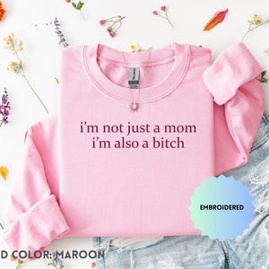 Könnte beinhalten: Ein hellrosa Sweatshirt mit dem gestickten Text "I'm not just a mom I'm also a bitch" in kastanienbraunem Faden. Das Sweatshirt wird mit Jeansshorts und floralen Akzenten präsentiert.