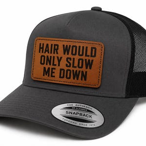 Op de afbeelding: Een antracietgrijze truckerpet met een zwarte mesh achterkant en een bruine rechthoekige patch aan de voorzijde. De patch bevat de zwarte tekst "HAIR WOULD ONLY SLOW ME DOWN". Een zilverkleurige en zwarte sticker op de klep vermeldt "THE AUTHENTIC YP CLASSICS SNAPBACK".