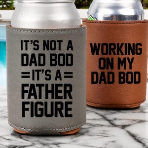 Op de afbeelding: Twee koelers voor drankblikjes. Eén is grijs met de tekst "IT'S NOT A DAD BOD = IT'S A = FATHER FIGURE." De andere is bruin met de tekst "WORKING ON MY DAD BOD."