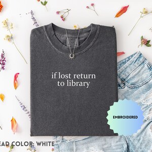 May include: Charcoal grey t-shirt with the white text "if lost return to library". The shirt is styled with denim shorts and a silver necklace. The image is decorated with colourful flowers and leaves.