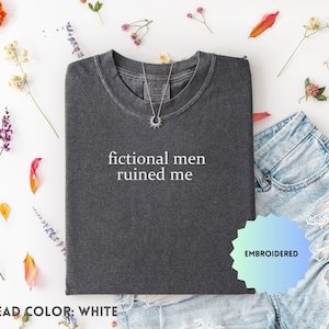 May include: Dark gray t-shirt with the phrase "fictional men ruined me" in white lettering. The shirt is paired with denim shorts and a silver necklace. The image is decorated with colorful flowers and leaves.