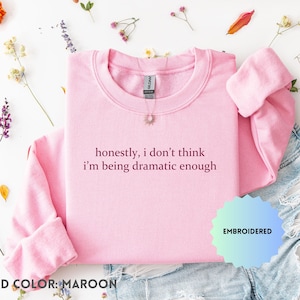 May include: A pale pink crew neck sweatshirt with the embroidered text "honestly, I don't think I'm being dramatic enough" in maroon thread. The sweatshirt is displayed with denim shorts and floral decorations.