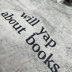 May include: A light gray sweatshirt with the phrase "will yap about books" embroidered in dark gray. The sweatshirt has a speckled pattern and a crew neck.