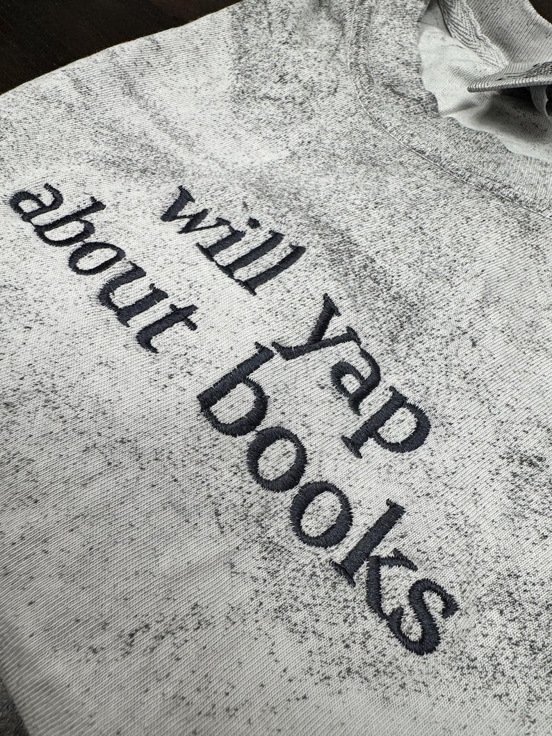 May include: A light gray sweatshirt with the phrase "will yap about books" embroidered in dark gray. The sweatshirt has a speckled pattern and a crew neck.