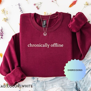 May include: A maroon crewneck sweatshirt with the phrase "chronically offline" in white. The sweatshirt has rolled-up sleeves and is paired with denim shorts. A silver necklace and floral accents complete the look. The thread color is white.