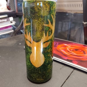 Puede incluir: Un vaso de acero inoxidable verde y dorado con un diseño de cabeza de ciervo de madera. El vaso tiene una tapa y es perfecto para mantener las bebidas frías o calientes.