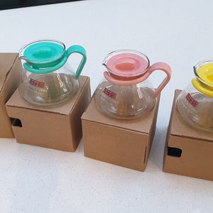 Puede incluir: Cuatro jarras de café de vidrio con tapas de diferentes colores: blanco, verde azulado, rosa y amarillo. Cada jarra está sentada en una caja de cartón marrón.