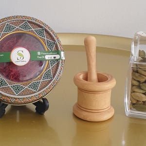 May include: A round tin of organic Persian saffron with a clear centre window, a small light-coloured wooden mortar and pestle, and a clear plastic container filled with green cardamom pods. The saffron tin features a green label with text.
