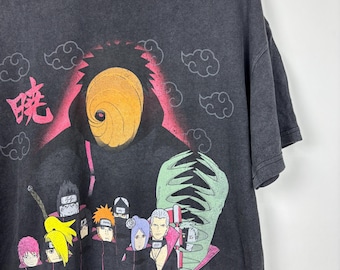 Puede incluir: Una camiseta gris carbón de manga corta con un estampado gráfico de varios personajes de anime con capas negras y nubes rojas. Una gran máscara naranja en espiral está centrada sobre los personajes, con texto japonés rosa a la izquierda.