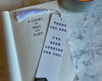 May include: A bookmark with the text "THERE YOU ARE. I'VE BEEN LOOKING FOR YOU" and a bat wing illustration. The bookmark is placed in a book titled "A COURT OF MIST AND FURY" with a lavender tassel. A lit candle and a second candle are in the background.