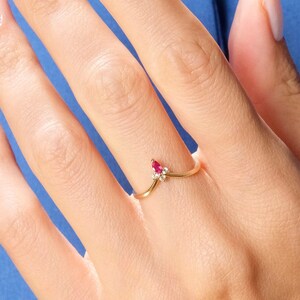 May include: A gold V-shaped ring featuring a small marquise-cut red gemstone and a tiny clear round gemstone. The ring is worn on a finger, and the text 1789 JEWELS is visible in the bottom right corner of the image against a blue background.