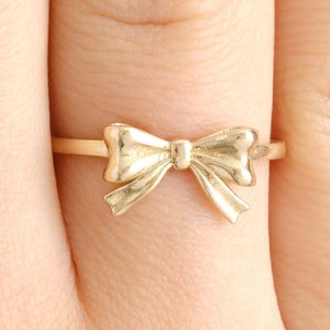 May include: A gold-coloured ring with a bow design. The bow is the central feature of the ring, with detailed, curved lines. The ring is displayed on a finger, against a soft background.