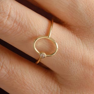 Puede incluir: Un delicado anillo de oro con una peque&ntilde;a gema transparente engastada en un dise&ntilde;o circular. El anillo tiene una banda delgada y elegante, con una est&eacute;tica minimalista. El anillo se muestra en el dedo de una persona.