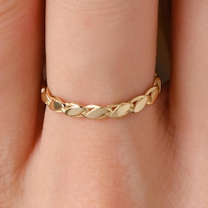 May include: A thin gold ring featuring a continuous, repeating leaf-like or braided pattern around the entire band. The metal has a polished, reflective finish that catches the light, creating a series of small, alternating highlights and shadows.