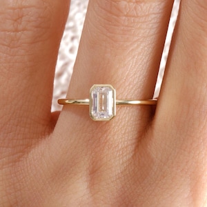 May include: A delicate gold ring featuring a rectangular, clear gemstone. The ring's band is thin and gold, complementing the bright, clear gemstone. The ring is worn on a finger, showcasing its elegant design. This ring is a piece of fine jewelry.