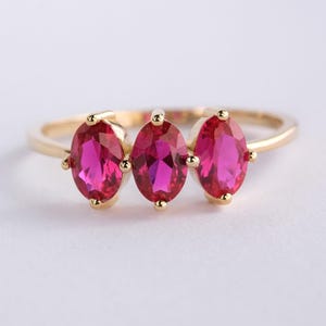 May include: A gold ring featuring three oval-cut, vibrant pink gemstones. The gemstones are set in a row across the top of the ring, with small gold accents on either side of each stone. The ring is displayed on a white background.