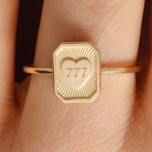 May include: A gold ring with a rectangular face featuring a heart and the number 777. The ring has a thin band and is shown on a finger. The face of the ring has a sunburst design around the heart.