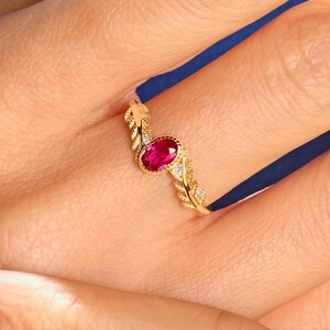 May include: A gold ring featuring a central oval-cut vibrant red gemstone. The band is designed with a textured, leaf-like pattern on both sides of the stone, accented with small, clear, sparkling stones embedded along the metal shank.