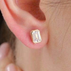 May include: A close-up of a gold stud earring with an emerald-cut, clear gemstone. The earring is set in a gold bezel and is worn on an ear. The earring is rectangular in shape.