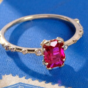May include: A gold ring featuring a rectangular, emerald-cut vibrant red gemstone set in a four-prong setting. The thin band is accented with small, round, clear stones embedded along the shank, which also displays a delicate beaded texture.