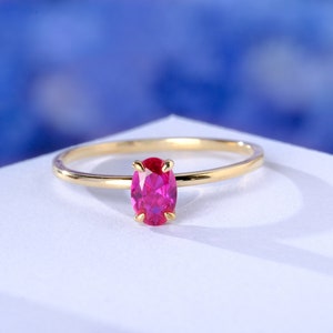 May include: A gold ring with a bright pink oval-shaped gemstone. The ring is on a white surface.