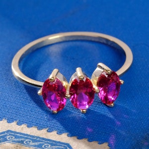 May include: A silver ring with three marquise-cut, vibrant pink gemstones. The ring has a delicate band and gold accents around the gemstones. The ring is displayed on a blue surface. The text "1789 JEWELS" is in the lower left corner.